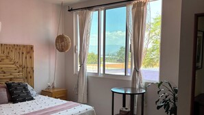 Une chambre à coucher comprenant un lit, un ventilateur de plafond, une fenêtre avec des rideaux, une table de chevet et une plante en pot.
