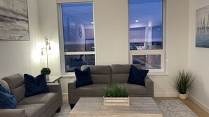 En stue med en beige sofa, en matchende lænestol og et sofabord med en plante. To vinduer med udsigt over byen, et billede på væggen og en gulvlampe.