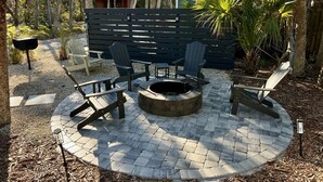 Un foyer circulaire, entouré de fauteuils Adirondack, avec une terrasse en bois et une clôture en bois en arrière-plan.