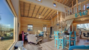 A spacious wooden cabin with a loft bedroom, a dining area, and a large window overlooking a scenic landscape. - 3 Chicks Ranch, 2 Cabins, Pool, Hot Tub & Hill Country (New Braunfels)
