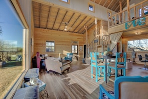 A spacious wooden cabin with a loft bedroom, a dining area, and a large window overlooking a scenic landscape. - 3 Chicks Ranch, 2 Cabins, Pool, Hot Tub & Hill Country (New Braunfels)