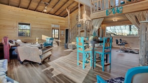 A spacious cabin interior with a high ceiling, wooden walls, a dining area with turquoise chairs, and a loft with a 'LOVE' sign. - 3 Chicks Ranch, 2 Cabins, Pool, Hot Tub & Hill Country (New Braunfels)