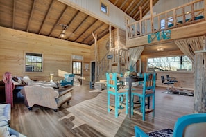 A spacious cabin interior with a high ceiling, wooden walls, a dining area with turquoise chairs, and a loft with a 'LOVE' sign. - 3 Chicks Ranch, 2 Cabins, Pool, Hot Tub & Hill Country (New Braunfels)