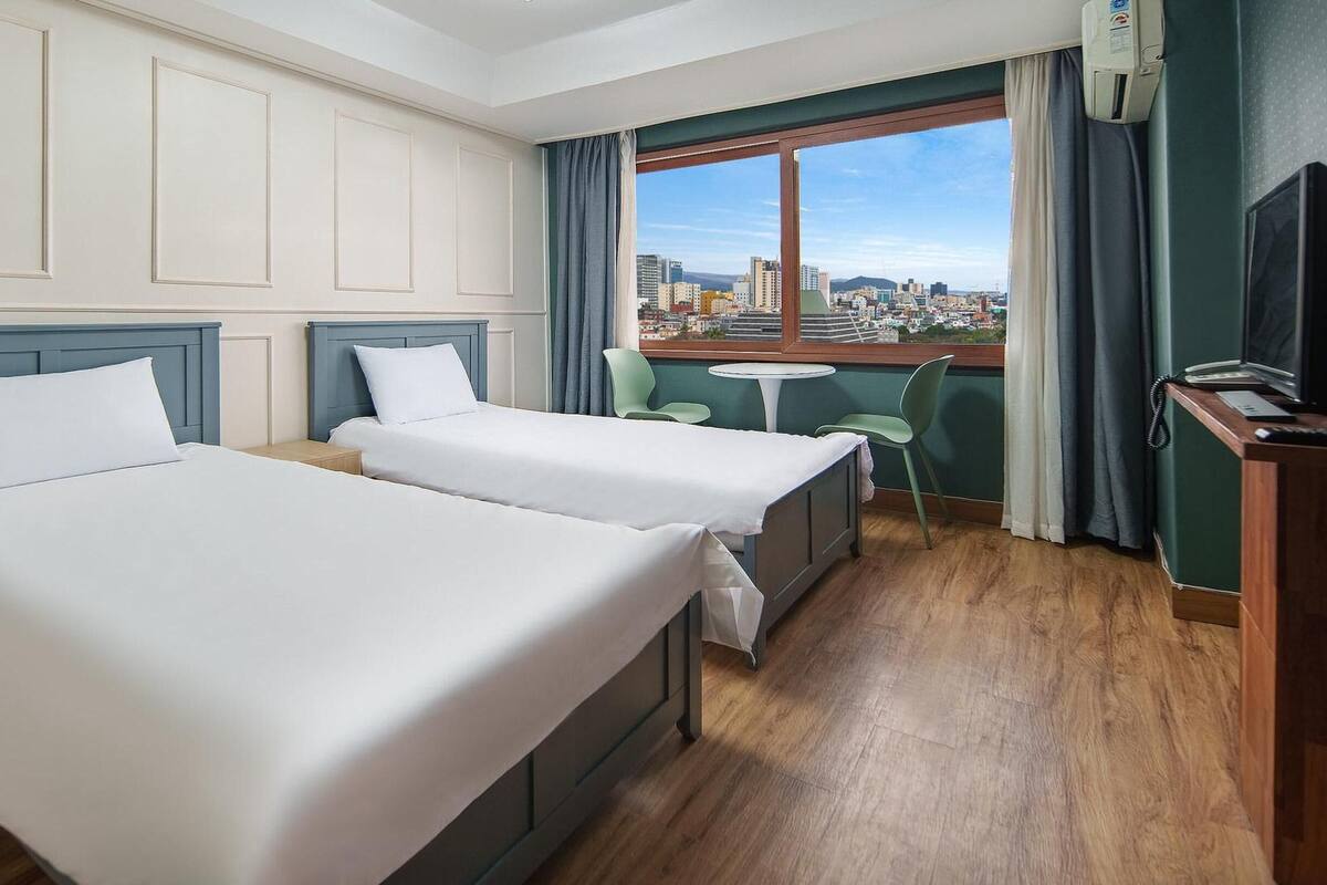 Ein Hotelzimmer mit zwei Betten, einem kleinen Tisch, Stühlen, einem Schreibtisch mit Telefon und einem Fenster mit Blick auf die Stadt.