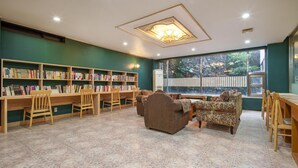 Een bibliotheek met boekenkasten, houten stoelen, een bank en een groot raam met uitzicht op groen.