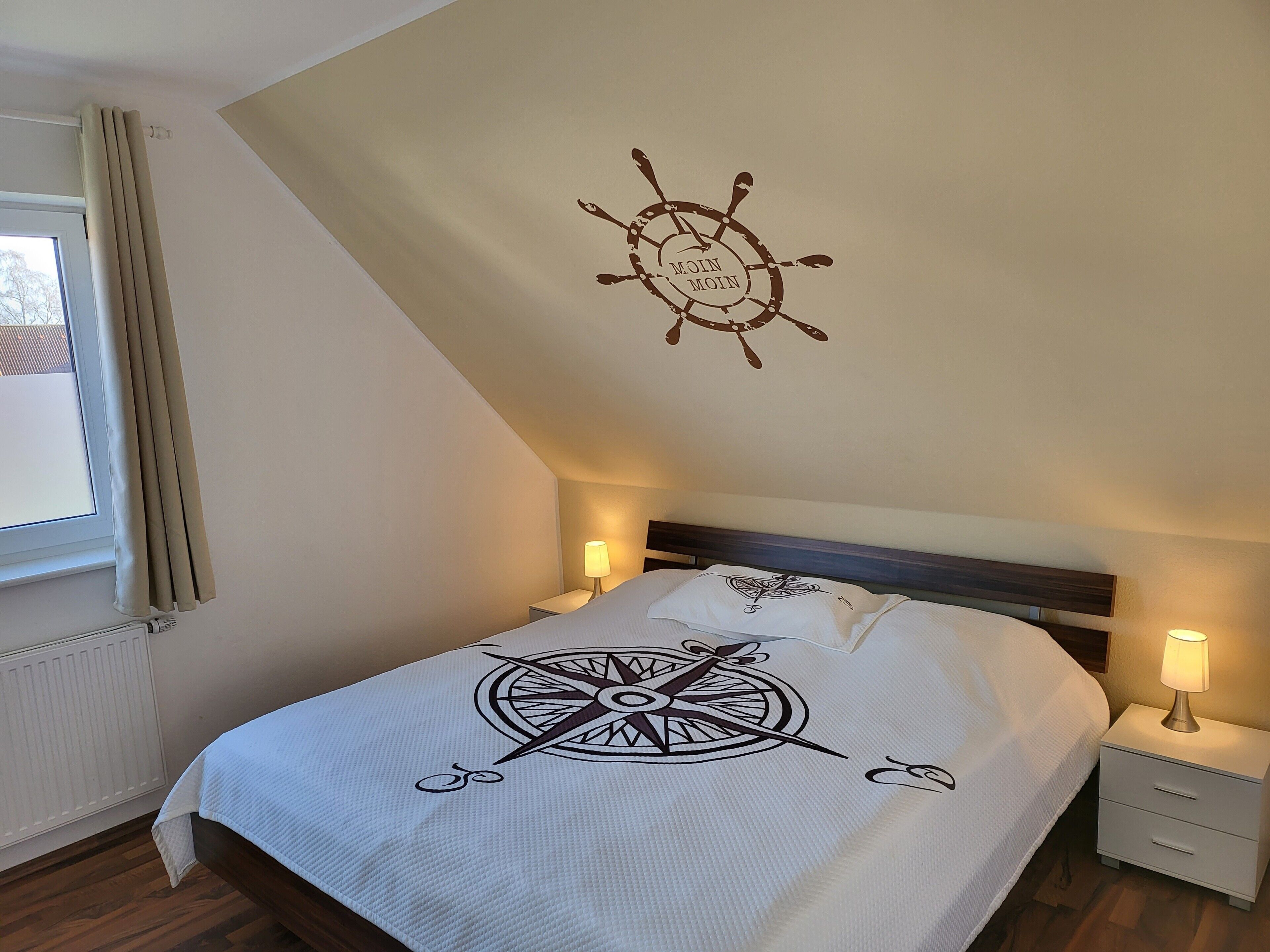 Ein Schlafzimmer mit einem Bett, einem Wandbild mit Kompassdesign, einem Fenster mit Vorhängen und einem Nachttisch mit Lampe.