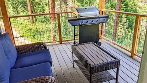 En overdækket terrasse med grill, fletmøbler og udsigt til et skovområde.