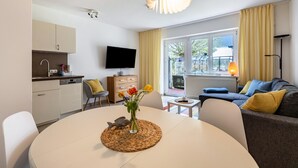 A modern living room with a sofa, a dining table with a vase of flowers, a kitchen area with cabinets and a TV, and a large window with curtains.