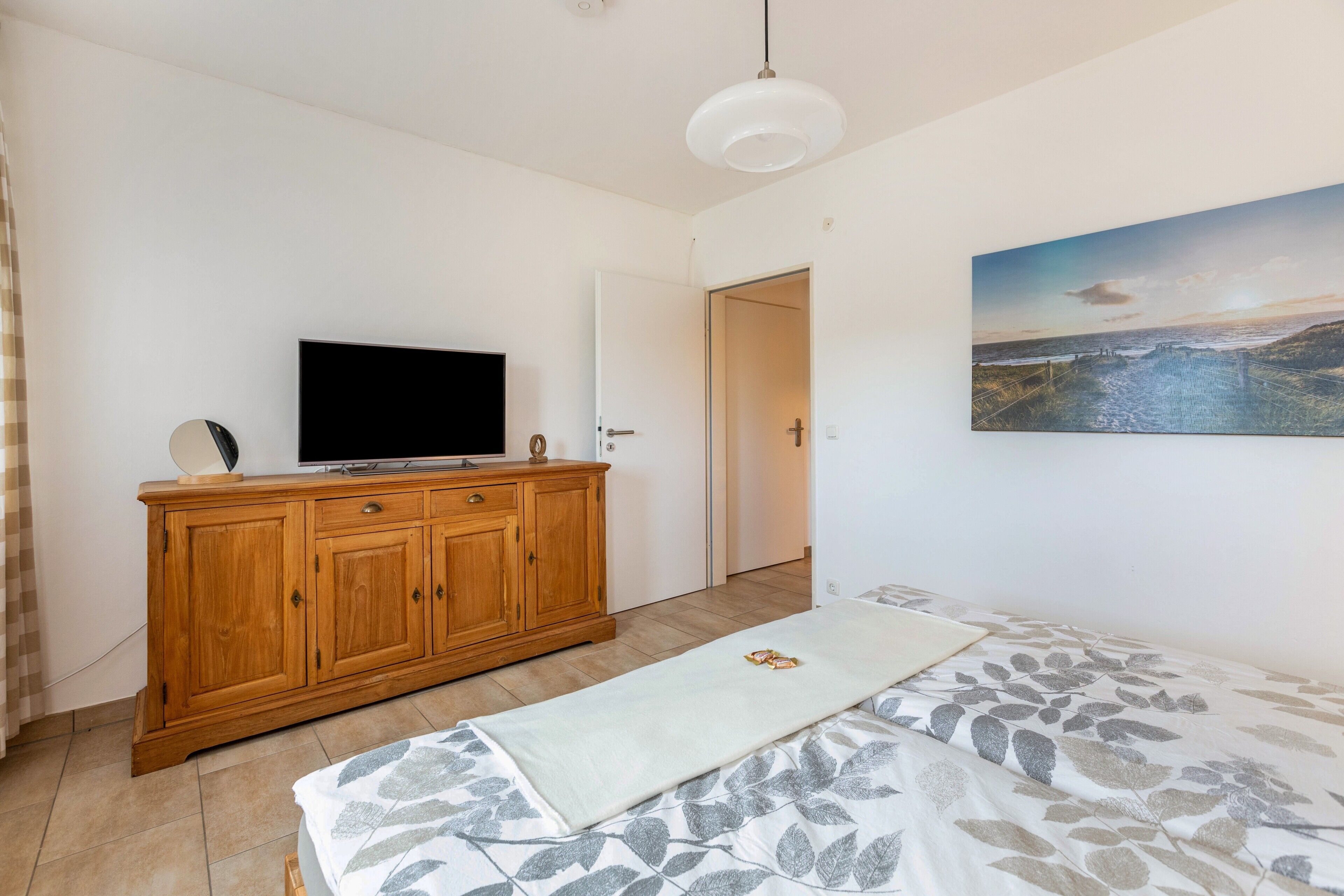 A bedroom with a bed, a TV on a wooden cabinet, a painting on the wall, and a door leading to another room.
