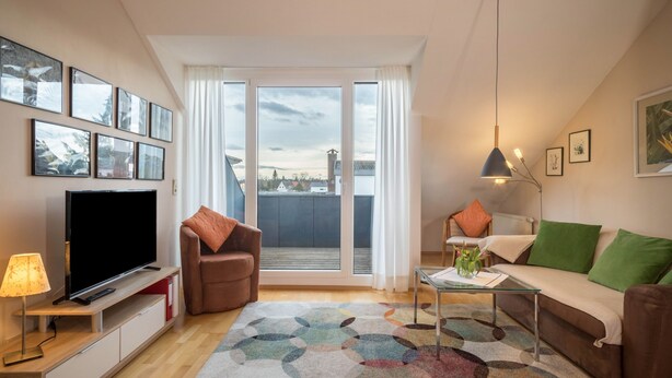 Ein modernes Wohnzimmer mit Flachbildfernseher, einer Couch, einem Sessel, einem Couchtisch und einem großen Fenster mit Blick.
