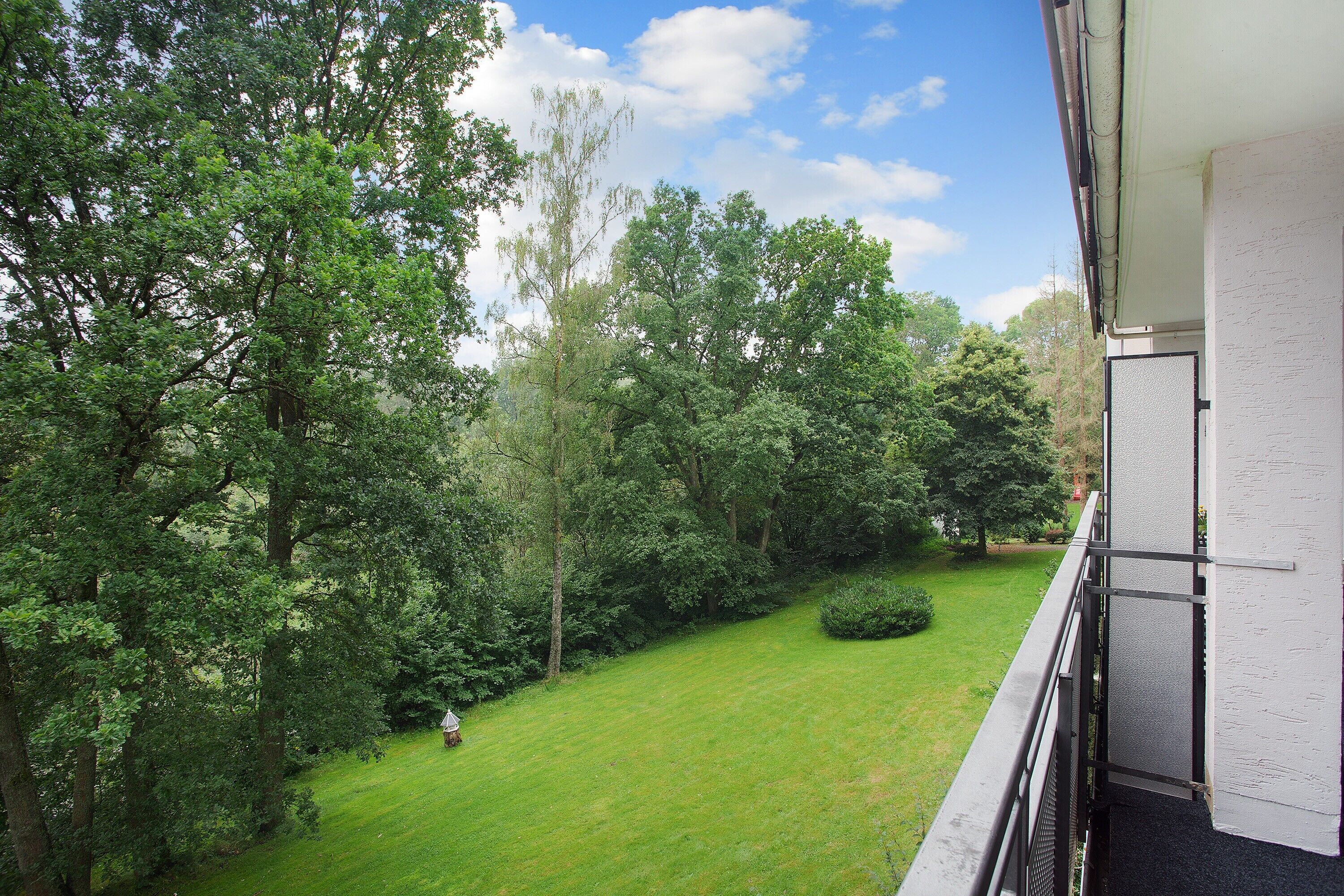 A balcony with a view of a well-maintained lawn and trees, a white wall, and a metal railing.
