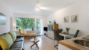 Un soggiorno moderno con divano, poltrone, un tavolino in vetro, zona pranzo con tavolo e sedie, cucina con lavello e ampie finestre vista alberi.