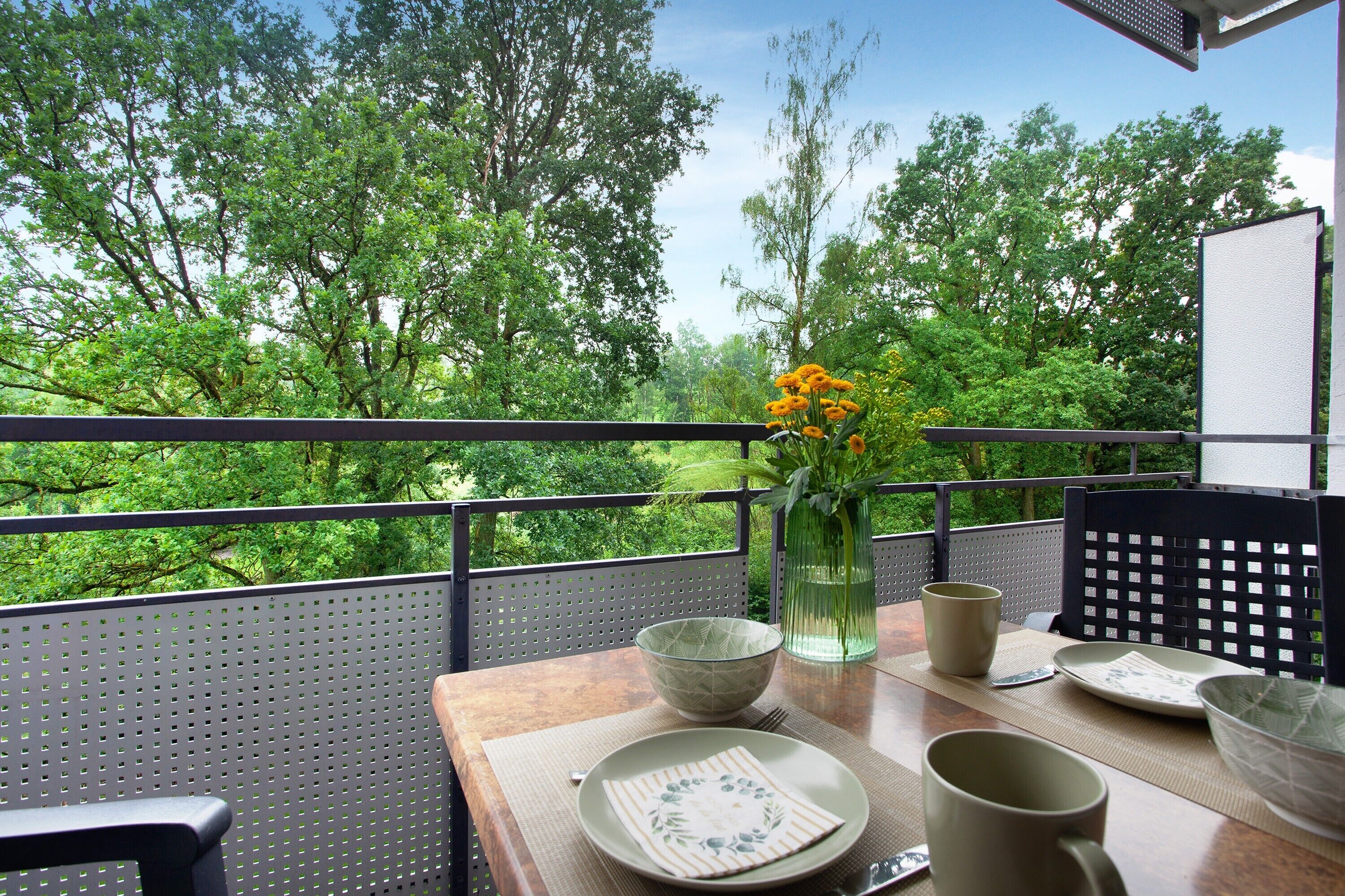 A balcony with a table set for dining, a vase with flowers, and a view of trees.