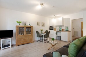 Ein modernes Wohnzimmer mit Flachbildfernseher, Holzschrank, Esstisch mit Stühlen und einer Küchenzeile mit weißen Schränken und Geräten.