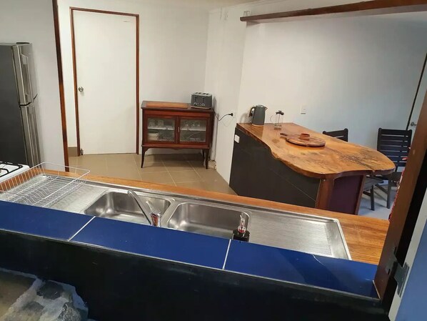 A compact kitchen with a sink, a stove, a wooden cabinet, and a dining area with chairs. - Treeverb Beach House : Hexagonal Beach house in the Daintree (Cow Bay)