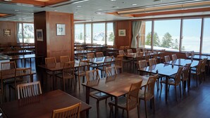 Uma sala de jantar ampla com várias mesas e cadeiras, grandes janelas com vista para as montanhas e painéis de madeira nas paredes.