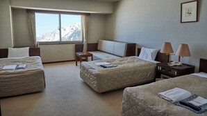 Quarto de hotel com duas camas, uma mesinha, uma mesa de cabeceira com abajur e uma janela com vista para as montanhas.