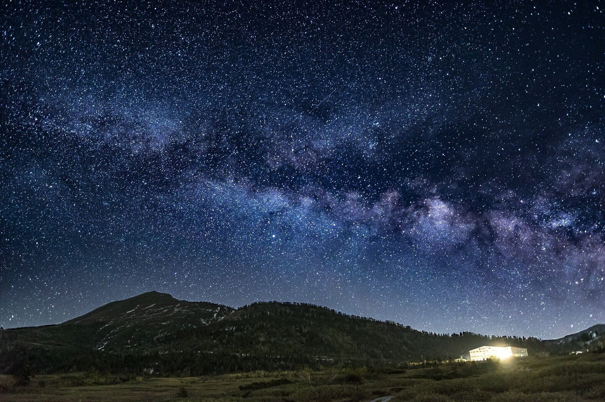 Cielo estrellado sobre un paisaje montañoso, con un edificio iluminado en primer plano.