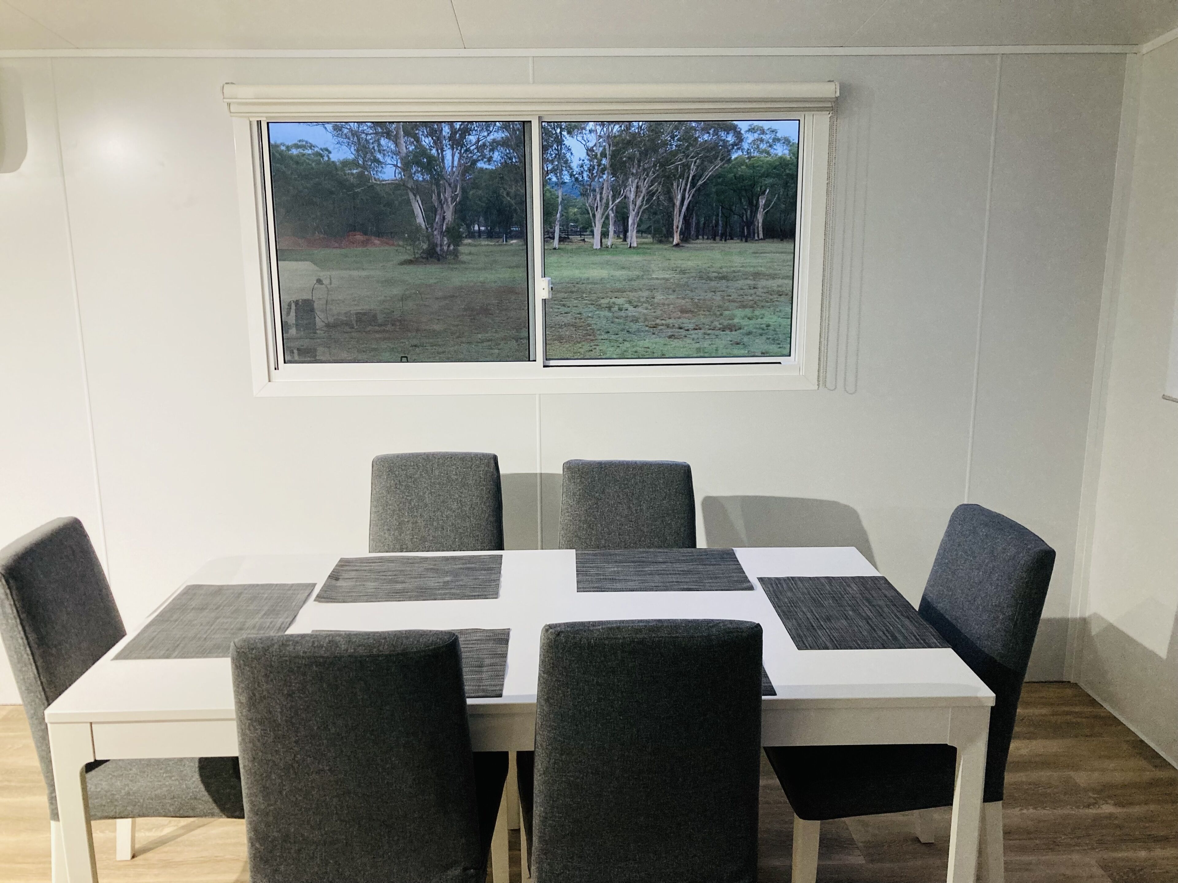 Ein Essbereich mit einem weißen Tisch und grau gepolsterten Stühlen, ein Fenster mit Blick auf Bäume und Gras sowie eine weiße Wand.