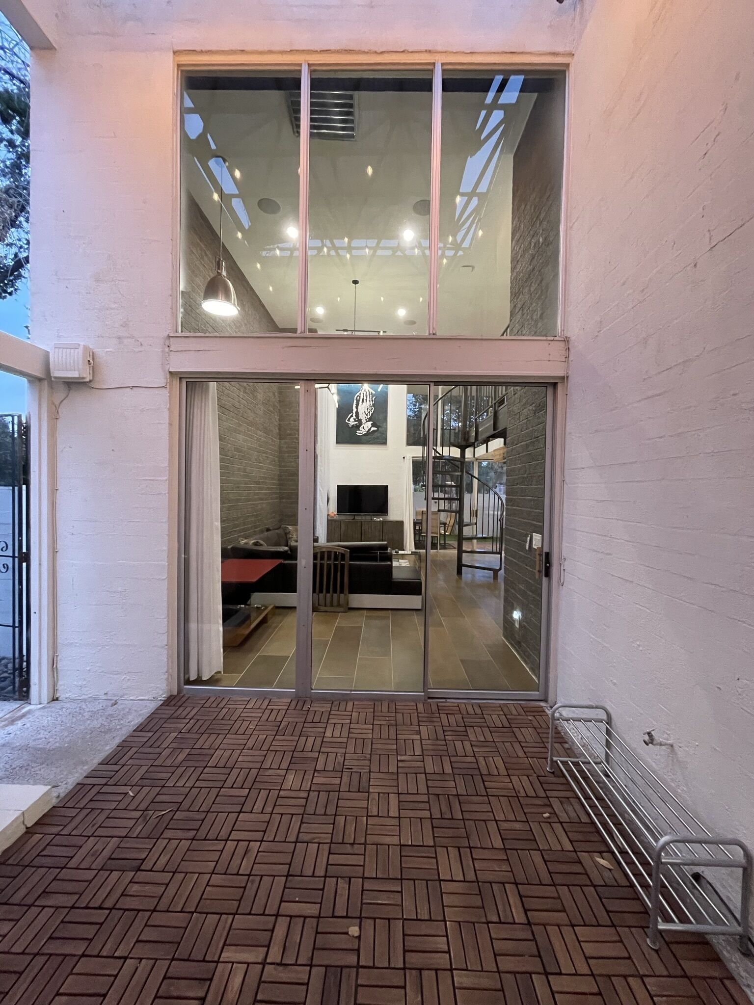 Ein modernes Gebäude mit Glastür, einer patioartigen Terrasse mit Ziegeloptik und einem einsehbaren Innenraum mit Treppe und Sitzbereich.