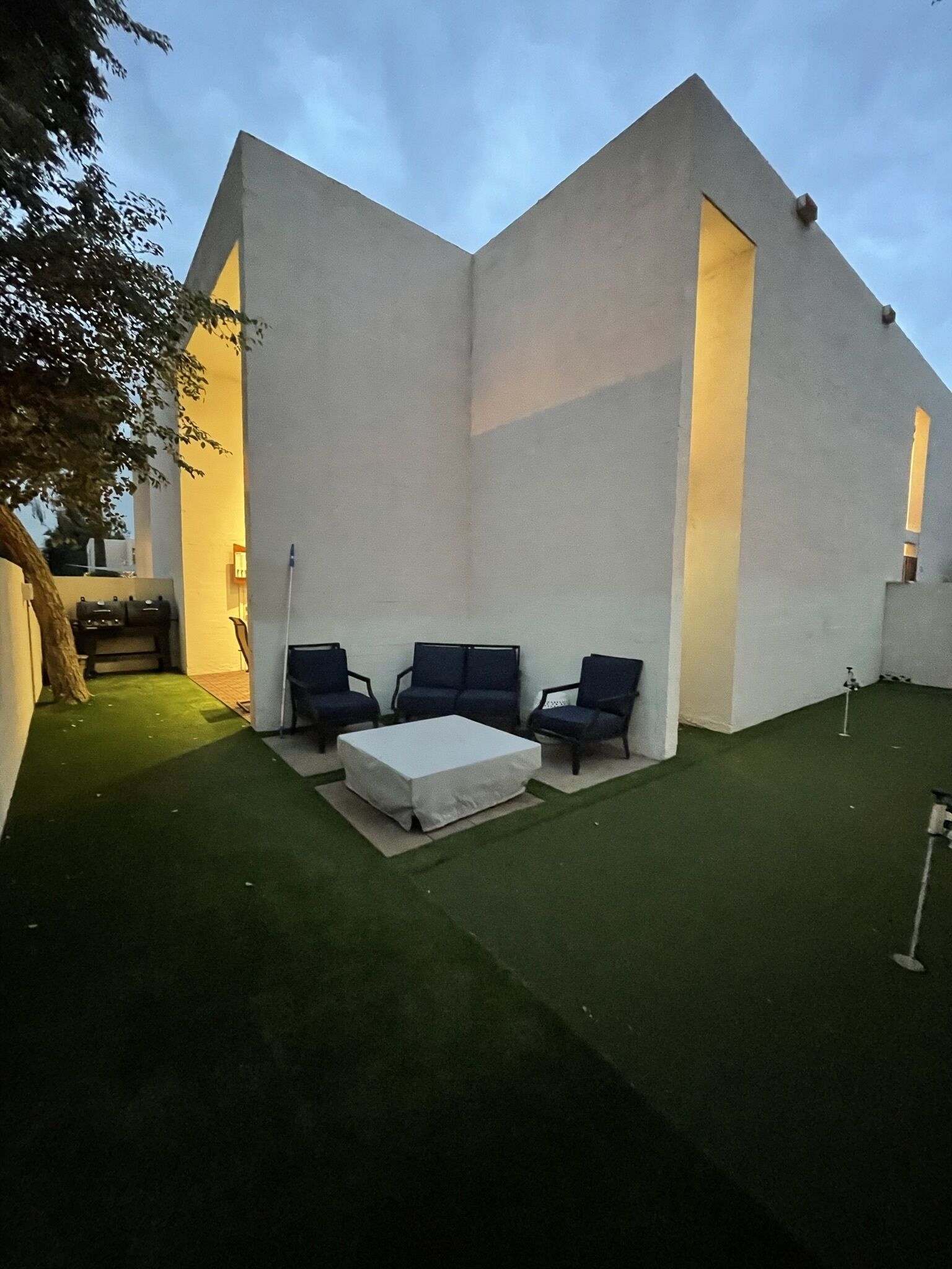 A modern building with a triangular facade, a small outdoor seating area with a white table and two chairs, and artificial green grass.