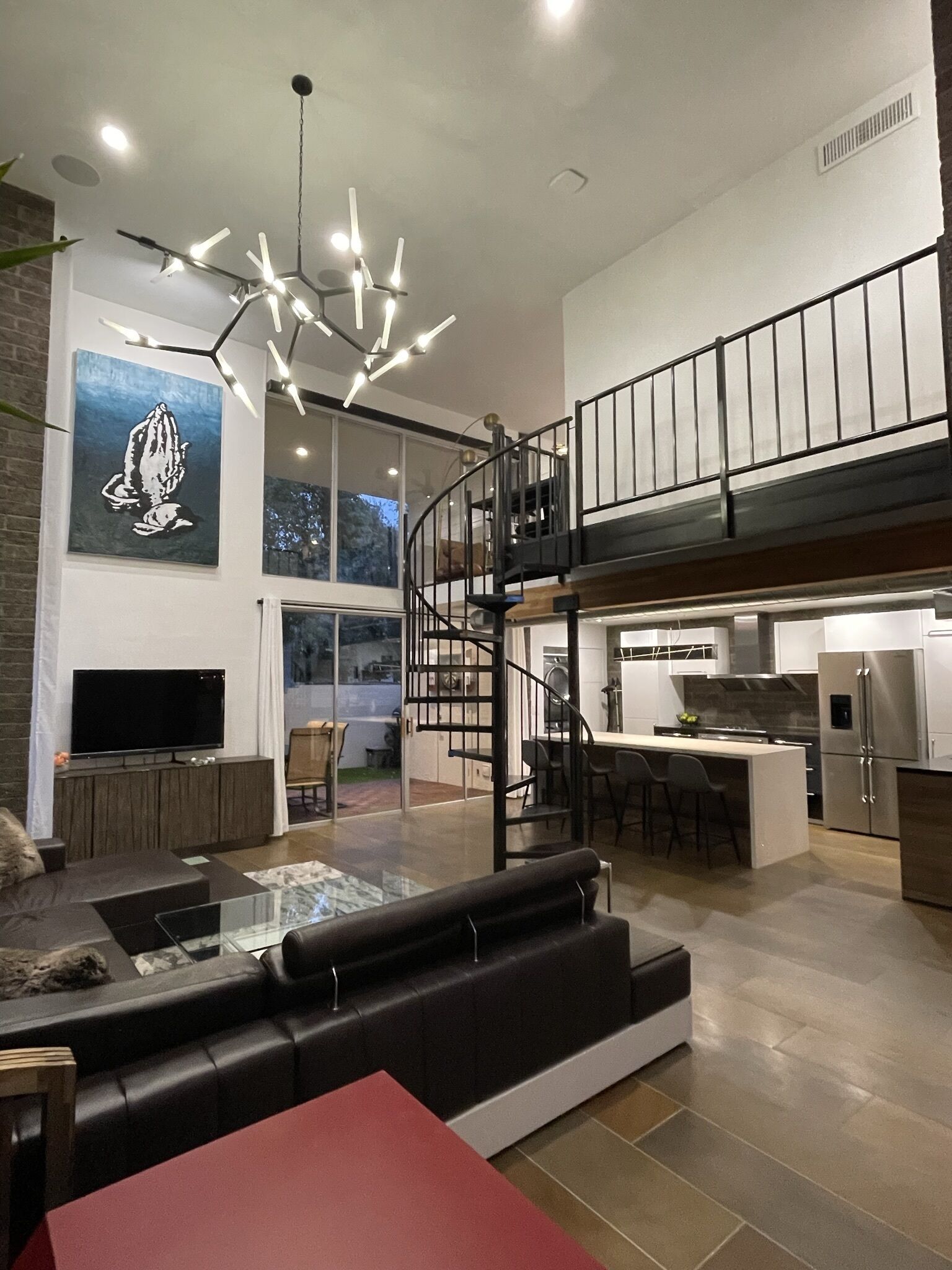 A modern living space with a black leather sofa, a glass coffee table, a red table, a TV, a kitchen with a stainless steel refrigerator, a staircase, and a large window.