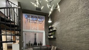 A modern living room with a sectional sofa, a glass coffee table, and a patterned rug. There is a staircase with a railing, a brick wall, and a ceiling light fixture. - 3b/2b "Beadle Box" Architectural Loft w/ Doghouse + Golf (Mesa)