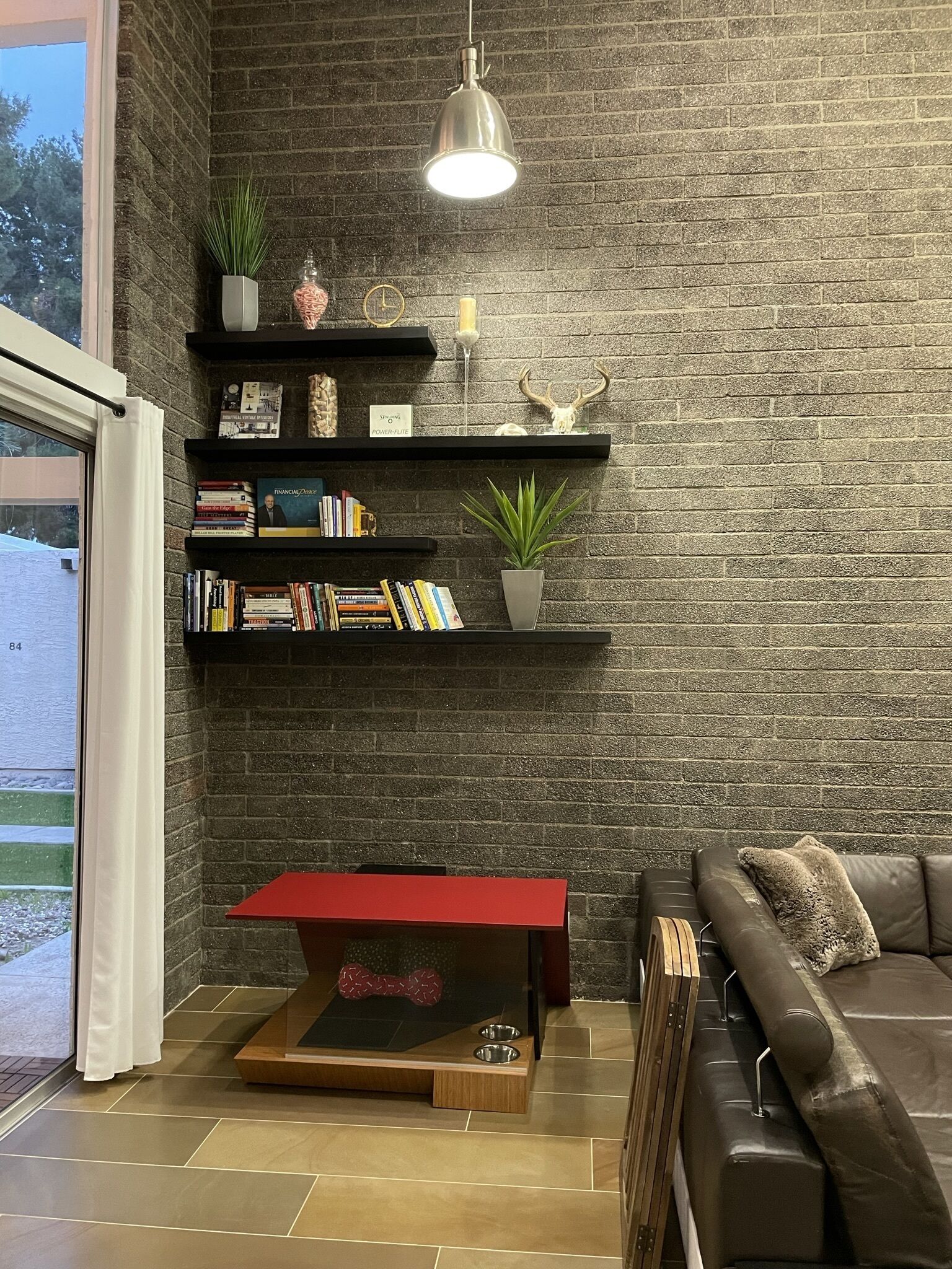 A modern living room with a brick wall, a red coffee table, a leather sofa, and a bookshelf with books and decorative items.
