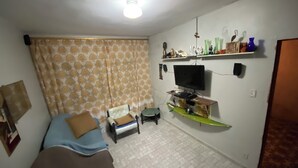 A small, well-lit room with a bed, a chair, a TV mounted on the wall, and a shelf with decorative items. - Casa de Praia 80 Metros do mar (Itanhaém)