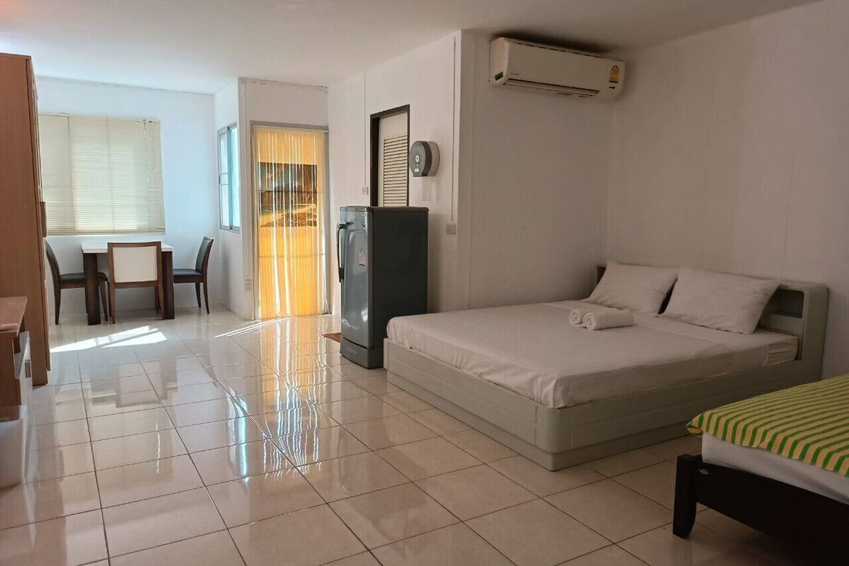 A hotel room with a bed, a desk, a chair, a refrigerator, and an air conditioning unit.