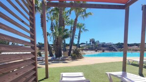 Un espacio al aire libre techado con césped artificial, dos sillones blancos y vista a una playa y palmeras.