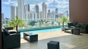 Terraza en la azotea con piscina, amueblada con mobiliario de mimbre y vistas a un paisaje urbano con edificios altos.