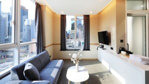 Habitación de hotel moderna con sofá, una mesita con un jarrón, televisor y kitchenette.