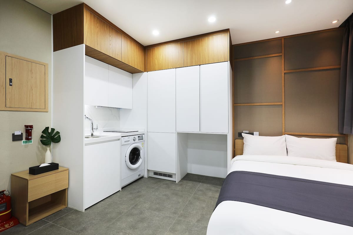 Een compacte hotelkamer met een bed, een klein keukenhoekje, een wasmachine en een houten kast.