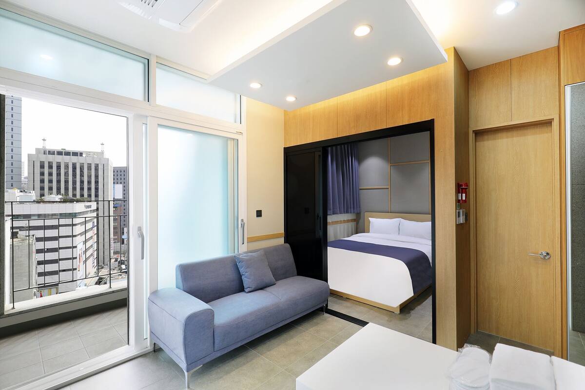 Een moderne hotelkamer met een bank, een bed met hoofdeinde, een houten garderobe en een groot raam met uitzicht op de stad.