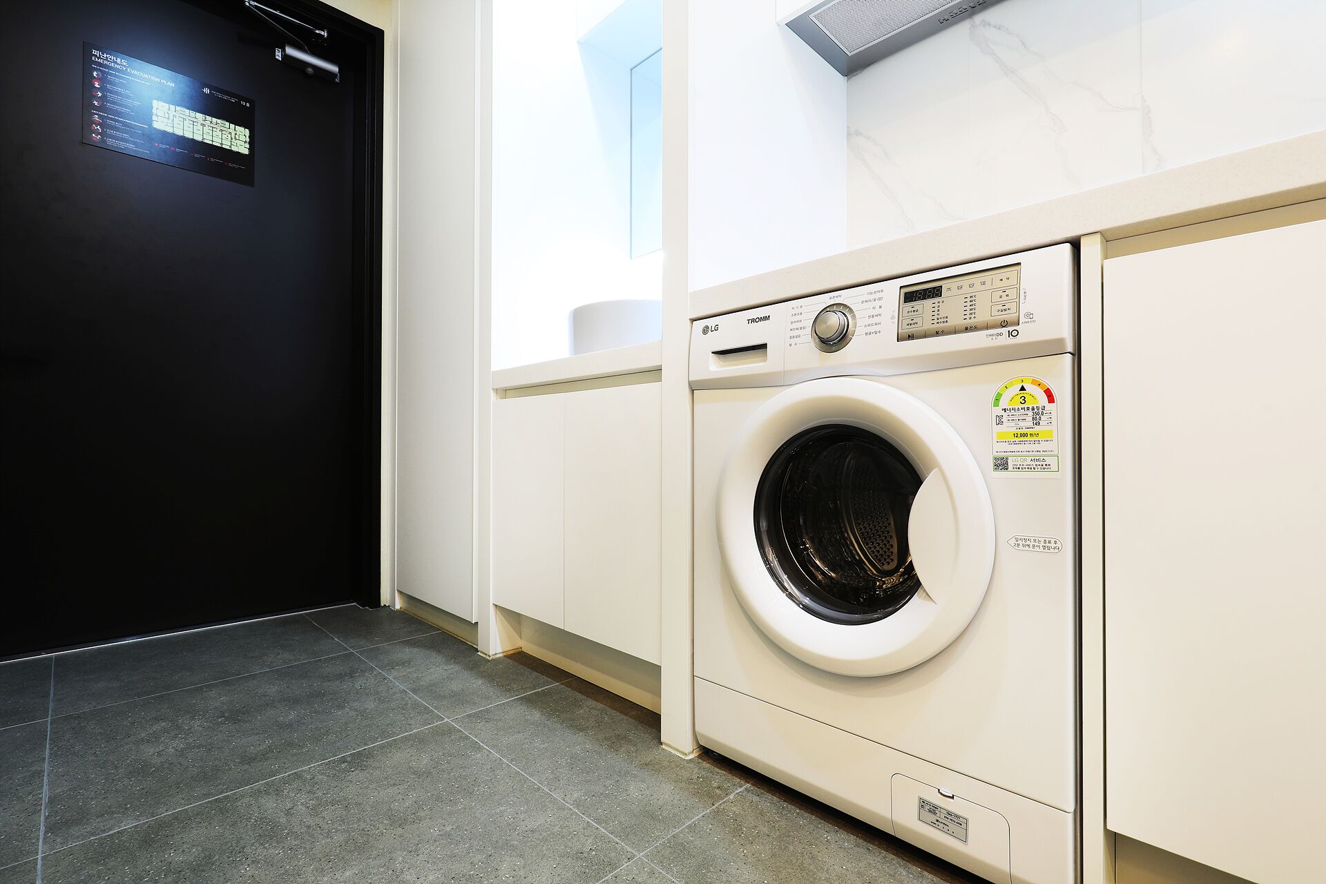 A laundry room with a front-loading washing machine, a black door, and a tiled floor.