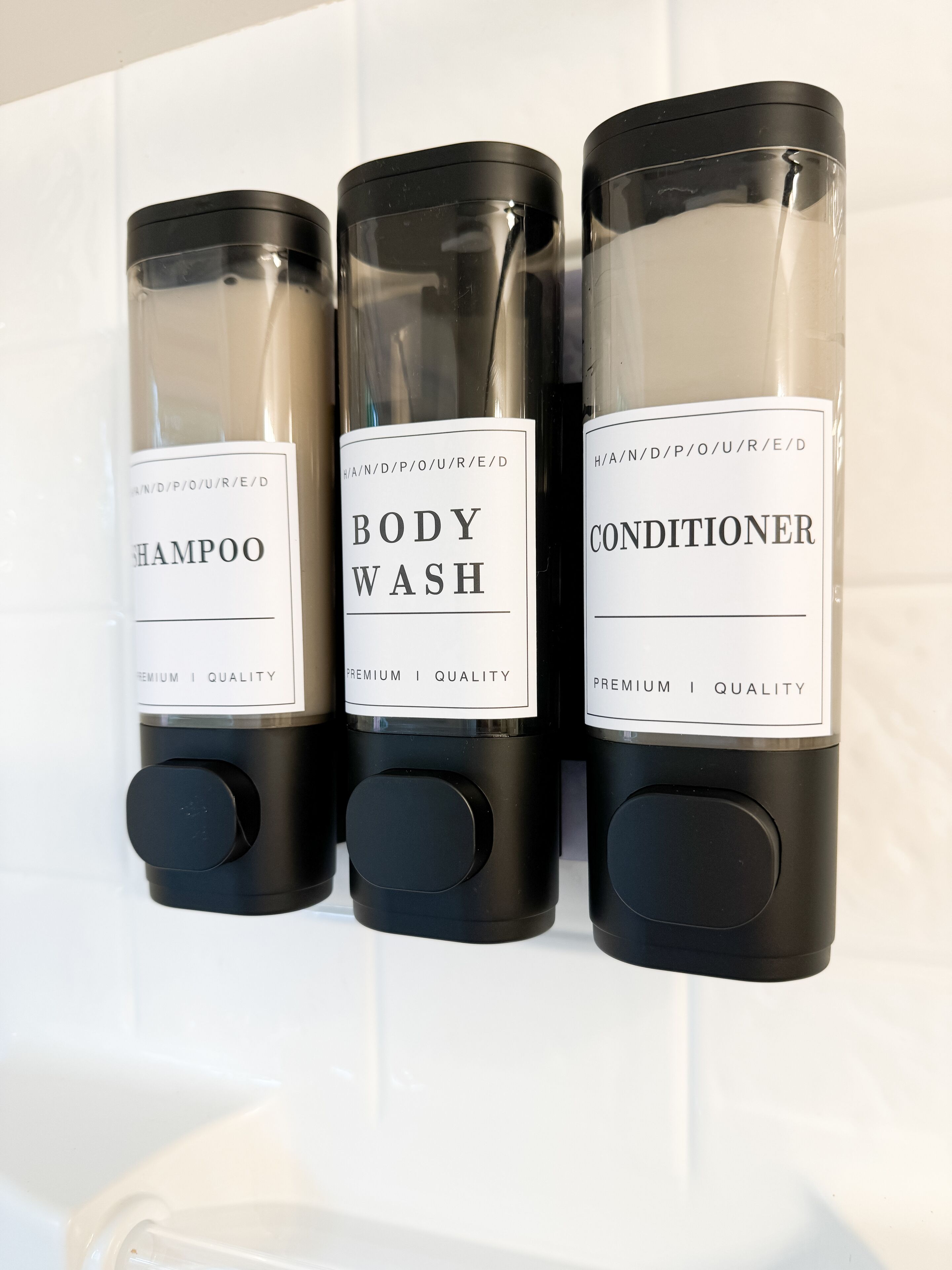 Three black dispensers with white labels for shampoo, body wash, and conditioner on a white tiled wall.