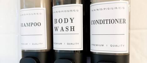 Three black dispensers with white labels for shampoo, body wash, and conditioner on a white tiled wall.