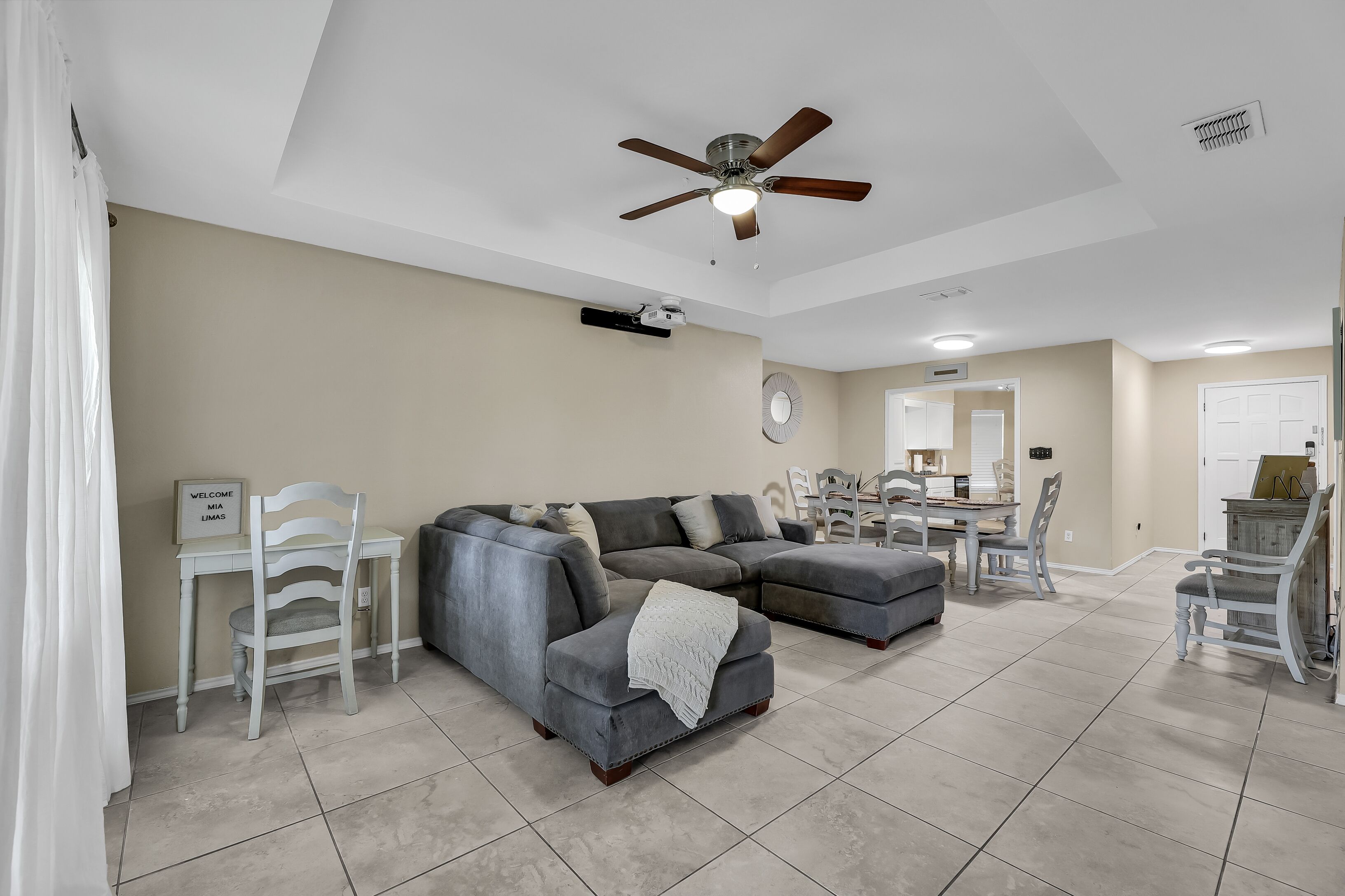 A living room with a grey sectional sofa, a ceiling fan, and a dining area with white chairs and a table.