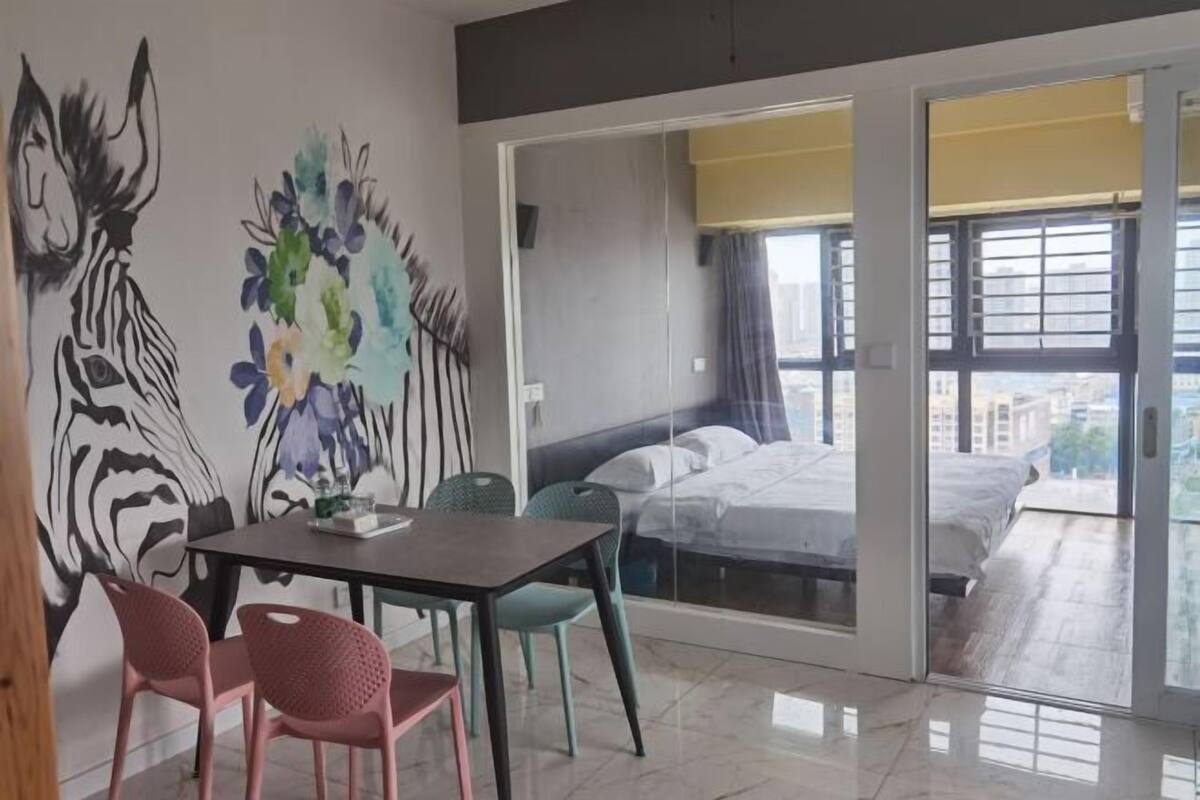 A modern room with a bed, a dining table with chairs, a large window, and a view of the city.