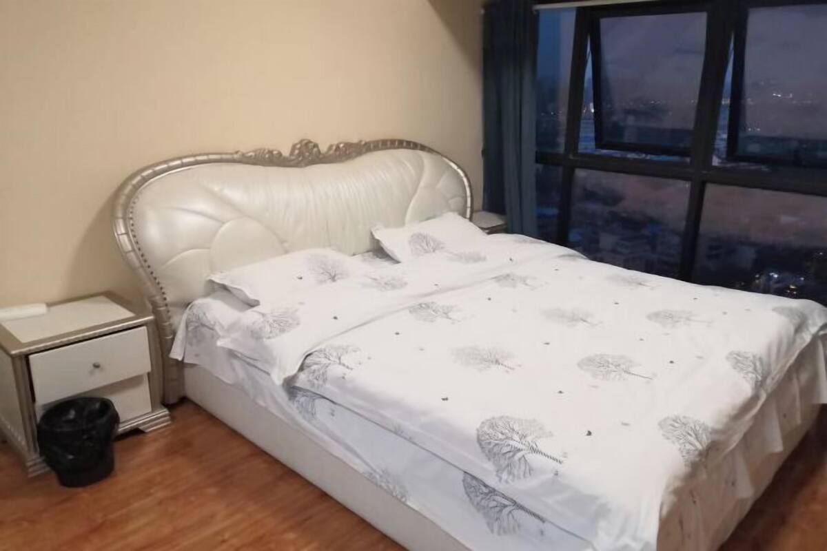 A bed with a white headboard, a nightstand with a drawer, and a black trash bin.