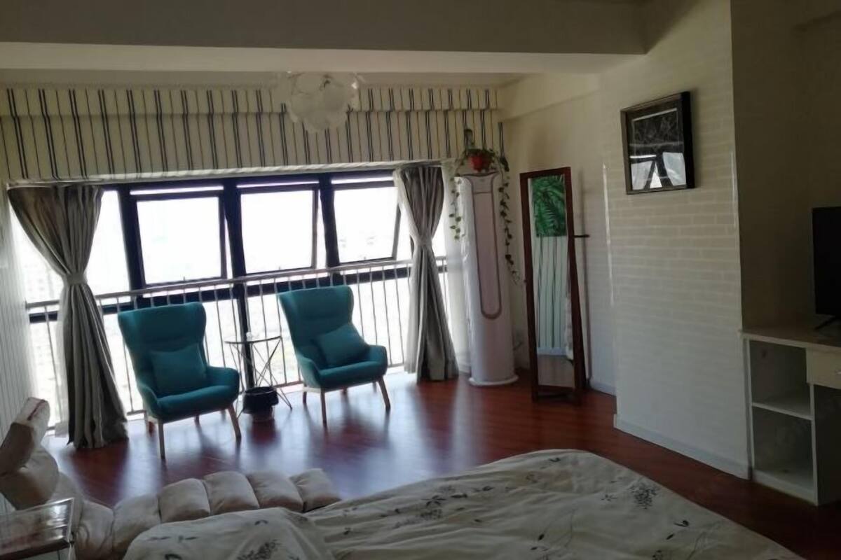 A hotel room with a bed, two chairs, a TV, a window with curtains, and a wall-mounted shelf.