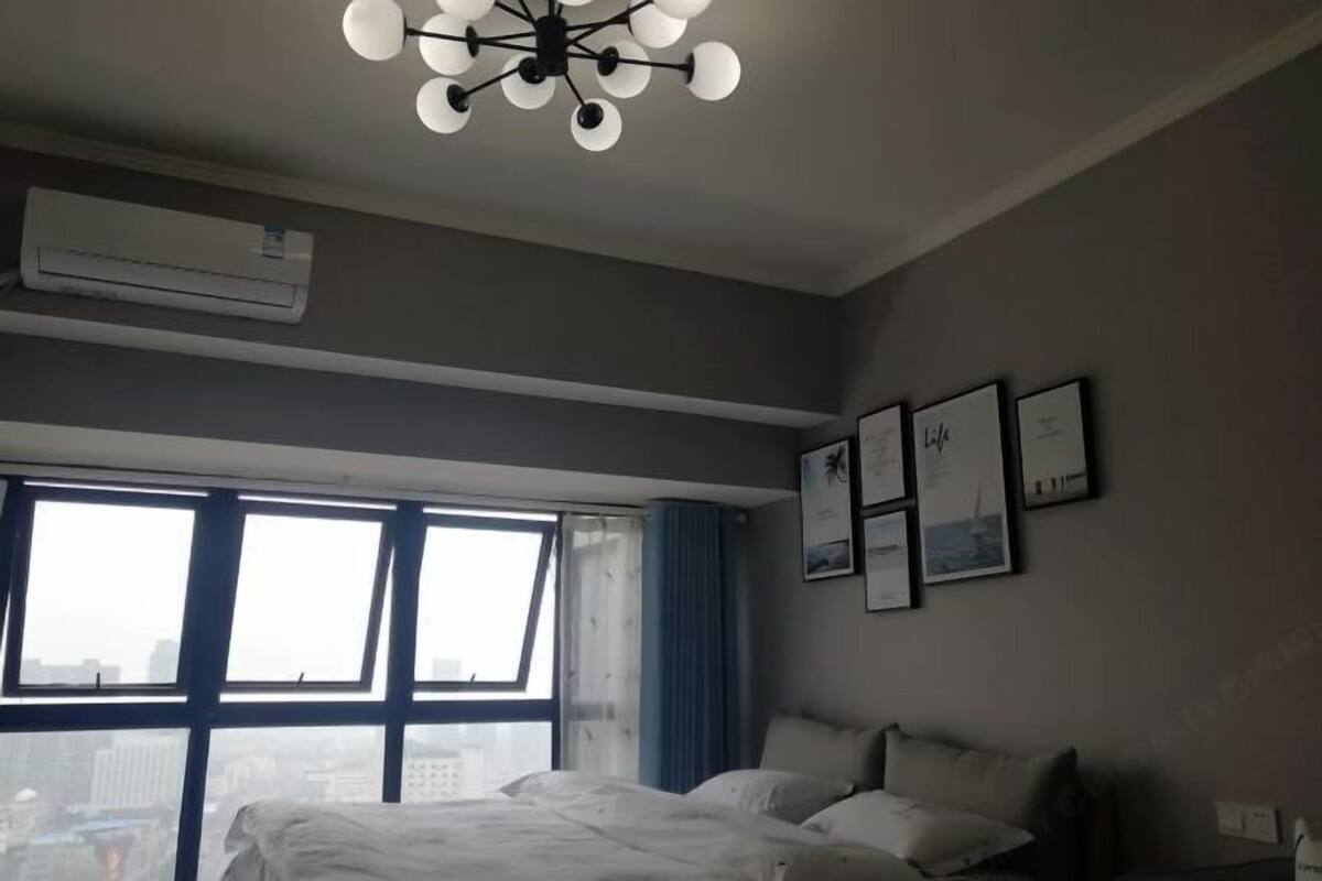 A hotel room with a large bed, a ceiling light fixture, an air conditioning unit, and a window with a city view.