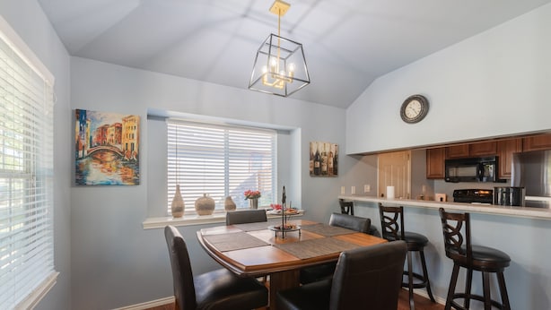 A dining area with a wooden table and chairs, a painting of a bridge, and a kitchen with wooden cabinets and a microwave.