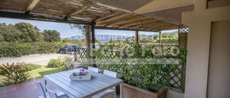 A covered patio with a table and chairs, a wooden pergola, and a view of a road and greenery.