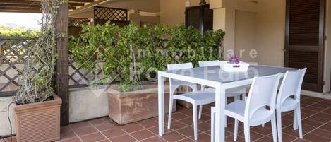 An outdoor patio with a white table and chairs, surrounded by potted plants and a wooden pergola.