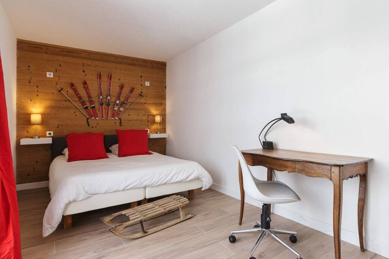A bedroom with a wooden headboard, a bed with red pillows, a desk with a chair, and skis mounted on the wall.