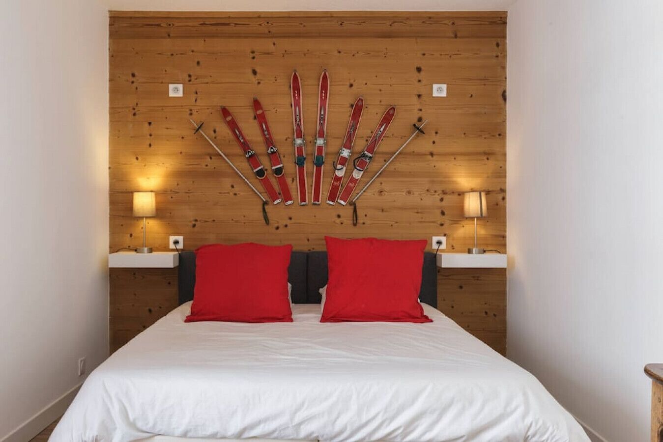 A bedroom with a wooden headboard, two red pillows, a white bedspread, and skis mounted on the wall.