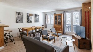 Ein geräumiges Wohnzimmer mit einem Holzessttisch, mehreren Stühlen, einer Couch und einem Fernseher. Große Fenster bieten Blick auf Bäume, und ein Balkon ist mit roten Geländern versehen.
