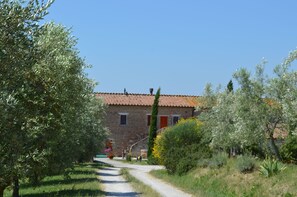Un bâtiment en pierre, doté d’un toit en tuiles, entouré d’oliviers et d’autres plantes.